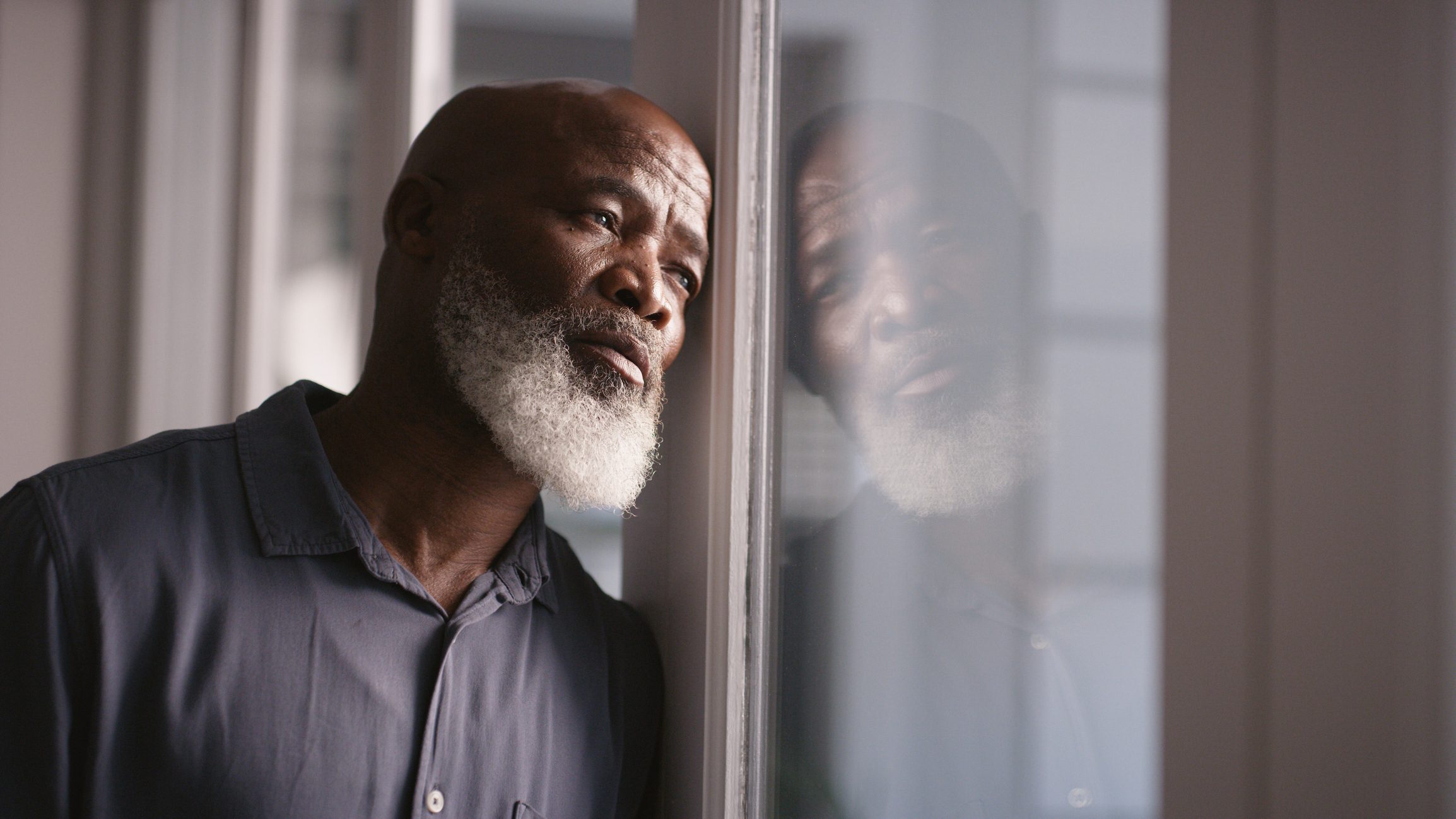 GettyImages-1452002001 Grieving man looks longingly out a window on a cloudy day.